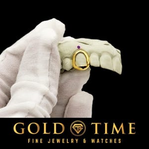 May include: A single gold tooth cap or grill piece fitted onto a white dental mold. The gold cap features a smooth, polished surface and is positioned over a central tooth. A small, circular purple gemstone is set into the dental mold above the cap.