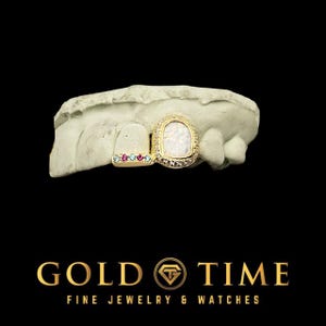 May include: A gold-colored grill with an opal-like center stone and small, colorful gems. The grill is displayed on a dental mold. The image also features the text "GOLD TIME FINE JEWELRY & WATCHES."