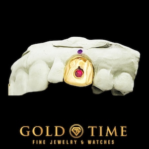 Puede incluir: Una tapa de diente de oro con una piedra preciosa roja engastada en un molde dental. Una pequeña piedra preciosa púrpura también está engastada en el molde. La imagen está sobre un fondo negro. El texto "GOLD TIME FINE JEWELRY & WATCHES" está en la parte inferior.
