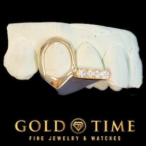 Op de afbeelding: Een goudkleurige tandgrill met vier kleine, heldere edelstenen. De grill wordt getoond op een witte tandafdruk tegen een zwarte achtergrond. De tekst "GOLD TIME FINE JEWELRY & WATCHES" staat onderaan.