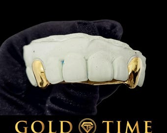 May include: A set of gold teeth grills fitted onto a white dental mould. The gold grill covers the two canine teeth and features a polished, reflective surface. The text GOLD TIME FINE JEWELRY & WATCHES is displayed at the bottom of the image.