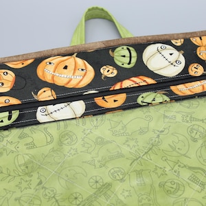 Może przedstawiać: Zielona i brązowa torba z zamkiem błyskawicznym i czarnym zamkiem. Torba jest ozdobiona wzorem pomarańczowych i białych dyń ze straszną twarzą. Wewnątrz torba jest wyłożona zielonym materiałem z białym wzorem kotów i gwiazd.