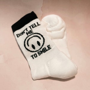 May include: A pair of white socks with a black cuff. The socks feature a black smiley face and the text "DON'T TELL ME TO SMILE" in black lettering. The socks are made of a soft, knitted material.