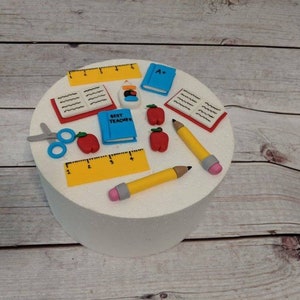 May include: A white, round cake decorated with school-themed fondant figures. Includes yellow pencils, rulers, red apples, blue books with the text "Best Teacher" and "A+", scissors, and open books.