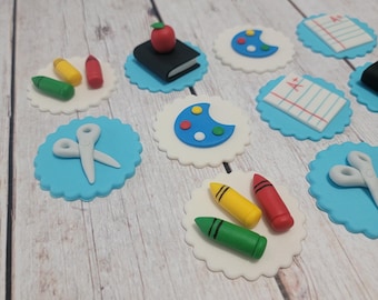 Puede incluir: Un conjunto de 10 adornos para cupcakes azules y blancos con varios diseños de temática escolar, que incluyen un libro con una manzana, una paleta de pintura, un par de tijeras, lápices de colores y una hoja de papel con una calificación de A+.