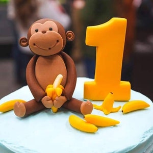 May include: A blue and white frosted cake with a brown monkey figurine holding a yellow banana. The cake is decorated with yellow banana slices and a yellow number one candle.