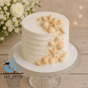 May include: A white frosted cake decorated with edible seashells, starfish, and pearls. The cake sits on a glass cake stand. The cake is a round, single-layer cake with a textured, ribbed frosting design.
