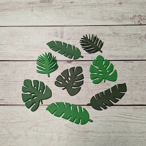 May include: A collection of nine green paper leaves in various shapes and sizes, including palm leaves and monstera leaves, arranged on a light wood surface.