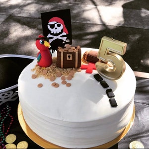 May include: A white cake decorated with a pirate theme. The cake has a red parrot, a treasure chest, a pirate flag, and a gold number 5 candle. The cake is on a gold plate.