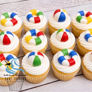 May include: A dozen cupcakes with white frosting, each topped with a colorful beach ball or life preserver. The cupcakes sit on a light wood-grain surface. The text "Carolina Cake Toppers" is visible in the lower left corner.