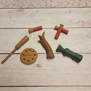 May include: A set of wooden toy hunting accessories, including a shotgun, a tree branch, a target, and two shotgun shells. The shotgun is green, the tree branch is brown, the target is brown with holes, and the shotgun shells are red and brown.