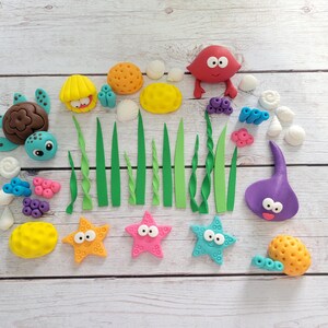 May include: A collection of colorful, handcrafted sea creature decorations, including a turtle, crab, starfish, and stingray. The set also features seaweed, coral, and shells, all arranged on a light wood surface.