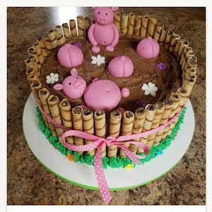 May include: A chocolate cake decorated with pink pig figurines and wafer cookies. The cake is surrounded by a fence made of wafer cookies and has a pink ribbon tied around it.