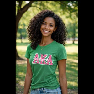 May include: A green v-neck t-shirt with the letters "AKA" in pink, outlined in white, and the words "alpha kappa alpha" below. The shirt is worn by a person with curly dark hair, smiling in an outdoor setting.
