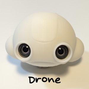 May include: A white drone with two black camera lenses. The drone has a rounded, smooth design.