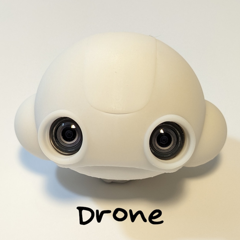 May include: A white drone with two black camera lenses. The drone has a rounded, smooth design.