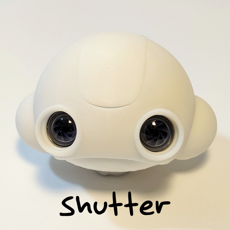 May include: A white, rounded, toy robot with two black lenses for eyes. The robot has a smooth, matte finish and a slightly raised, rounded top. The word "Shutter" is written in black below the robot.