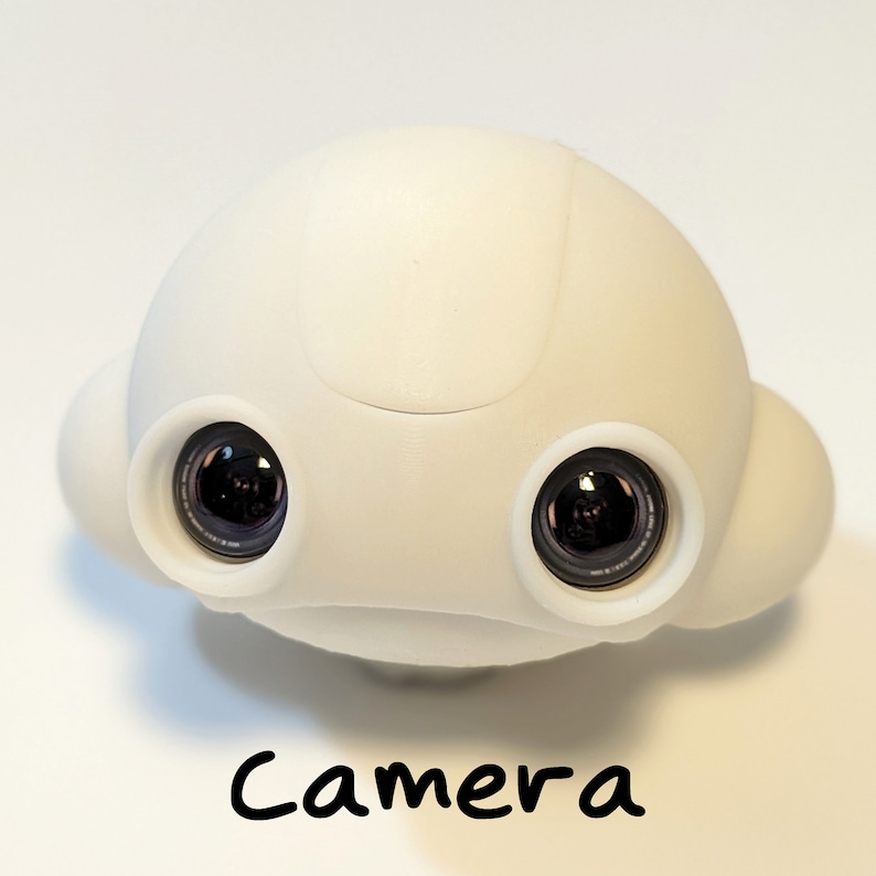 May include: A white, rounded, toy robot with two black camera lenses. The robot has a smooth, plastic surface and a slightly curved top. The word "Camera" is written below the robot.