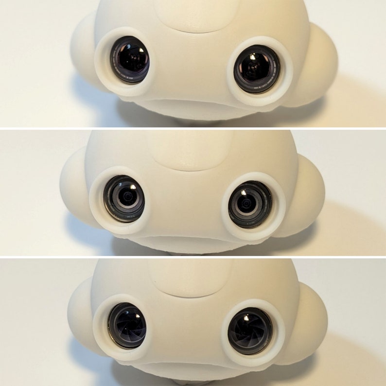 May include: A white robot head with two black camera lenses. The robot head has a rounded shape with two small ear-like protrusions on the sides.