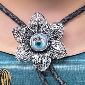 May include: A silver-colored bolo tie featuring a flower-shaped pendant with an eye-shaped center. The eye has a blue iris and a black braided cord. The pendant is made of a silver-colored metal with an openwork design.