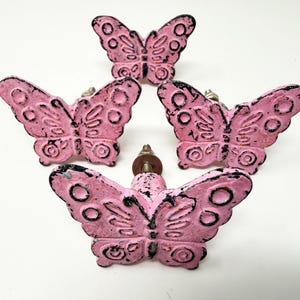 May include: A set of four butterfly-shaped drawer knobs in a distressed pink finish with black accents. Each knob features a textured, raised wing pattern and a central metal screw post for attachment, showcasing a weathered, vintage-inspired look.