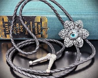 May include: A silver-toned bolo tie with a flower-shaped pendant featuring a blue eye design. The pendant is intricately detailed. The braided cord is dark gray, and the tie has silver-toned tips. The background includes a book with the text "TALES Edgar Allan Poe".