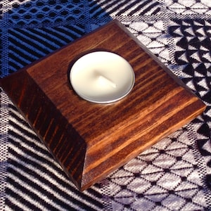 May include: A square wooden candle holder with a dark brown finish. A white tea light candle is placed in the center of the holder.