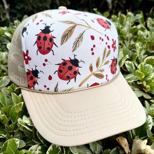 May include: A beige trucker hat with a white front panel decorated with ladybugs and floral motifs. The hat has a curved peak and a mesh back. The ladybugs are red with black spots, and the flowers are red and gold.