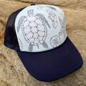 May include: A navy blue trucker hat with a light blue front panel featuring a sea turtle and other sea life illustrations. The hat has a navy blue brim and a black mesh back.