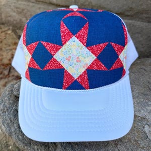May include: A white trucker hat with a quilted design on the front. The design features a blue background with red star shapes and a floral center. The brim is white and the mesh backing is also white.