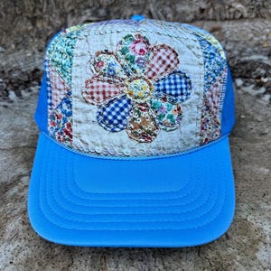 May include: A bright blue baseball cap featuring a front panel decorated with a quilted fabric patch. The patch displays a large, multi-colored floral design made from various patterned fabrics, including gingham and small floral prints, with stitching.