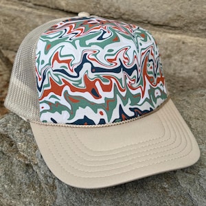 May include: A beige trucker hat with a mesh back and a curved brim. The front panel has a white background with a colourful abstract pattern in green, orange, and navy blue. A tan rope detail accents the brim.