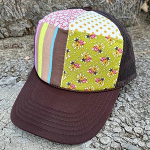 Op de afbeelding: Een bruine truckerpet met een patchwork-ontwerp. Het patchwork bevat roze bloemenpatronen, witte stippen en groene bloemenpatronen. De klep is effen bruin en de mesh-achterkant is donkerbruin.