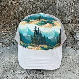 May include: A white trucker hat with a landscape design. The front panel features a scenic mountain and forest illustration in shades of green, blue, and orange. The hat has a mesh back and a curved brim.