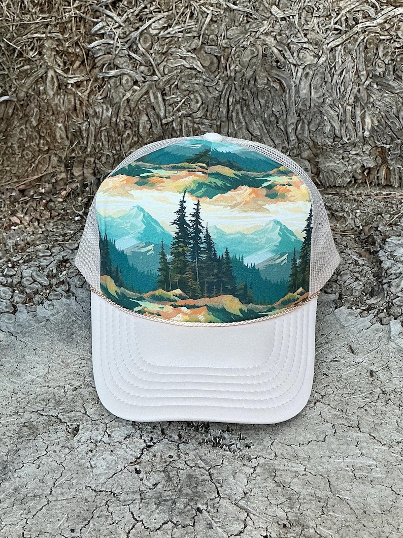 May include: A white trucker hat with a landscape design. The front panel features a scenic mountain and forest illustration in shades of green, blue, and orange. The hat has a mesh back and a curved brim.