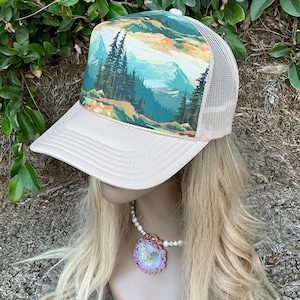 May include: A beige trucker hat with a mesh back and a landscape design on the front panel. The landscape features mountains, trees, and a colorful sky. The hat has a curved brim and a snap closure.