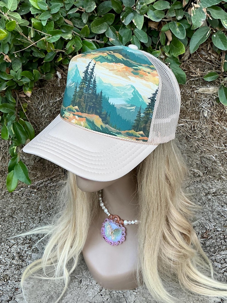 May include: A beige trucker hat with a mesh back and a landscape design on the front panel. The landscape features mountains, trees, and a colorful sky. The hat has a curved brim and a snap closure.