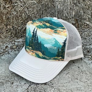 May include: A white trucker hat with a landscape design. The front panel features a scenic view of mountains, trees, and a colorful sky. The hat has a white brim and a beige mesh back.