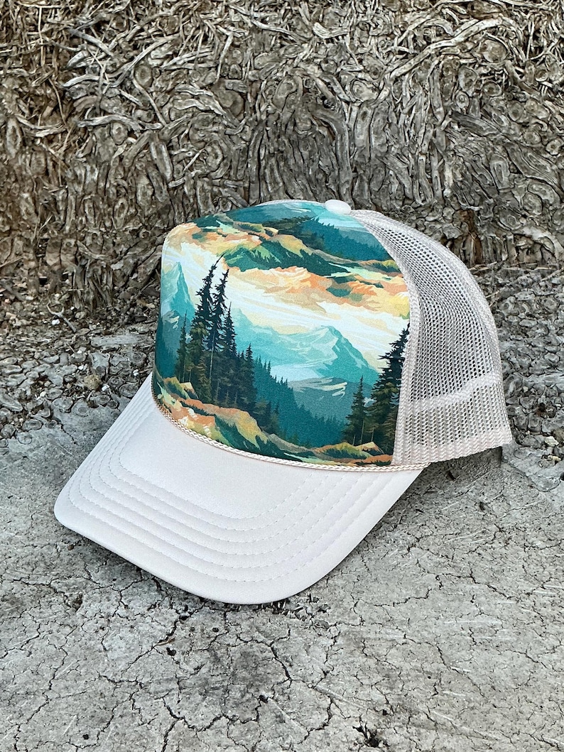 May include: A white trucker hat with a landscape design. The front panel features a scenic view of mountains, trees, and a colorful sky. The hat has a white brim and a beige mesh back.