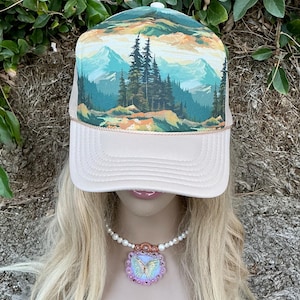 May include: A trucker hat with a landscape design featuring mountains and trees. The hat has a cream-colored front panel and a mesh back. A necklace with a butterfly pendant is also visible.