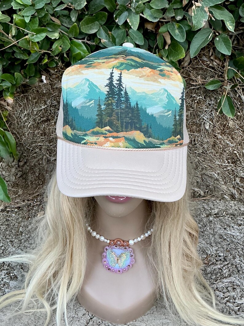 May include: A trucker hat with a landscape design featuring mountains and trees. The hat has a cream-colored front panel and a mesh back. A necklace with a butterfly pendant is also visible.