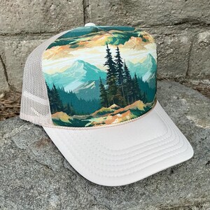 May include: A white trucker hat with a mesh back and a landscape design. The front panel features a colorful illustration of mountains, trees, and a river. The hat has a curved brim and a beige rope detail.