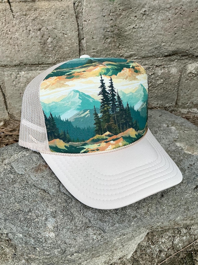 May include: A white trucker hat with a mesh back and a landscape design. The front panel features a colorful illustration of mountains, trees, and a river. The hat has a curved brim and a beige rope detail.