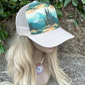May include: A trucker hat with a white front panel and a beige mesh back. The hat features a scenic landscape design with mountains, trees, and a colorful sky. The brim is white, and the hat is displayed on a mannequin head.
