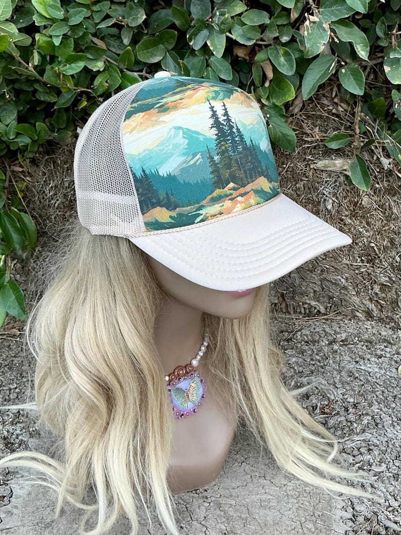 May include: A trucker hat with a white front panel and a beige mesh back. The hat features a scenic landscape design with mountains, trees, and a colorful sky. The brim is white, and the hat is displayed on a mannequin head.