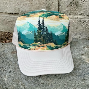 May include: A trucker hat with a landscape design. The front panel features a mountain scene with pine trees, mountains, and a colorful sky. The brim is white, and the mesh backing is tan.