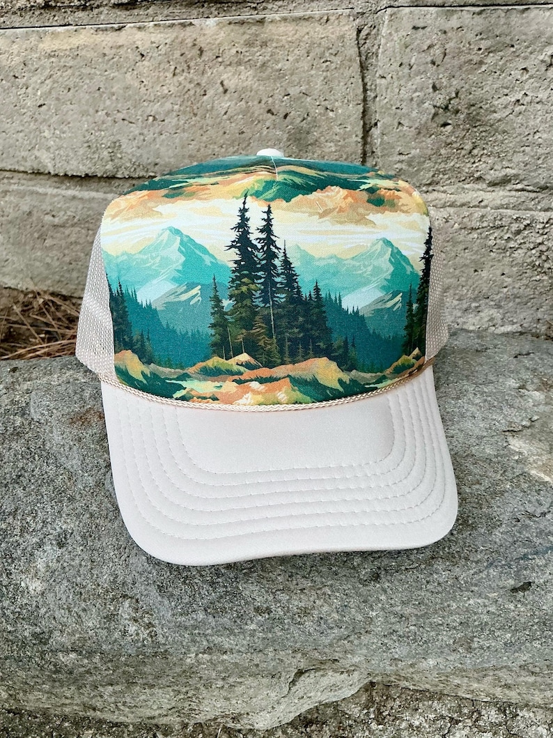 May include: A trucker hat with a landscape design. The front panel features a mountain scene with pine trees, mountains, and a colorful sky. The brim is white, and the mesh backing is tan.