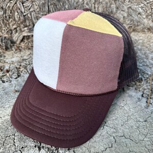 May include: A patchwork trucker hat with a brown brim and a white front panel. The top of the hat features a color block design with pink, yellow, and mauve panels. The mesh back is dark brown.