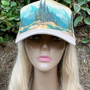 May include: A beige trucker hat with a scenic mountain landscape design. The hat features a curved brim and a mesh back. The artwork depicts a forest, mountains, and a blue sky. The hat is a stylish accessory for outdoor enthusiasts.