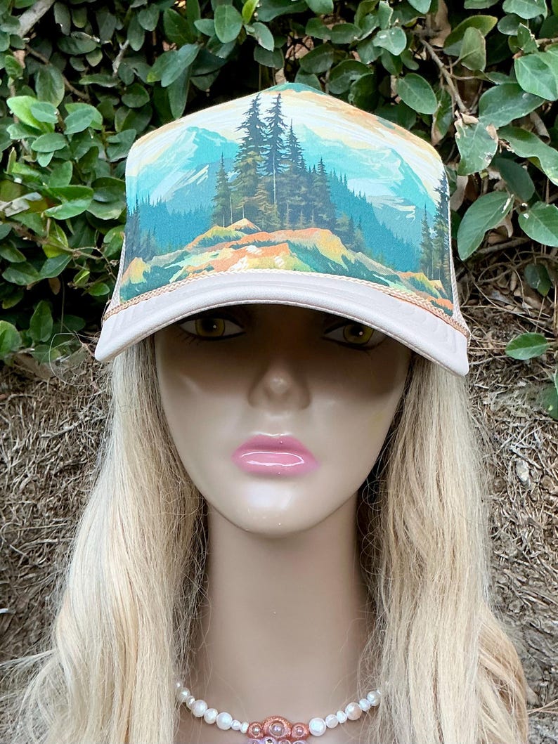 May include: A beige trucker hat with a scenic mountain landscape design. The hat features a curved brim and a mesh back. The artwork depicts a forest, mountains, and a blue sky. The hat is a stylish accessory for outdoor enthusiasts.